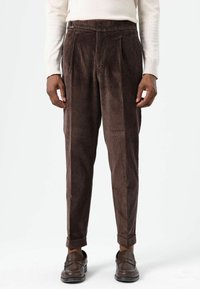 Pantalon en velours côtelé marron avec plis, coupe fuselée, ourlets retroussés et texture lisse, associé à des mocassins foncés.