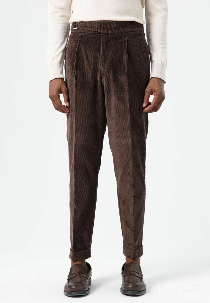 Pantalon en velours côtelé marron avec plis, coupe fuselée, ourlets retroussés et texture lisse, associé à des mocassins foncés.
