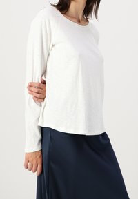 Camicetta bianca a maniche lunghe realizzata in tessuto morbido e leggero, con vestibilità rilassata e scollatura rotonda, abbinata a una gonna blu scuro.