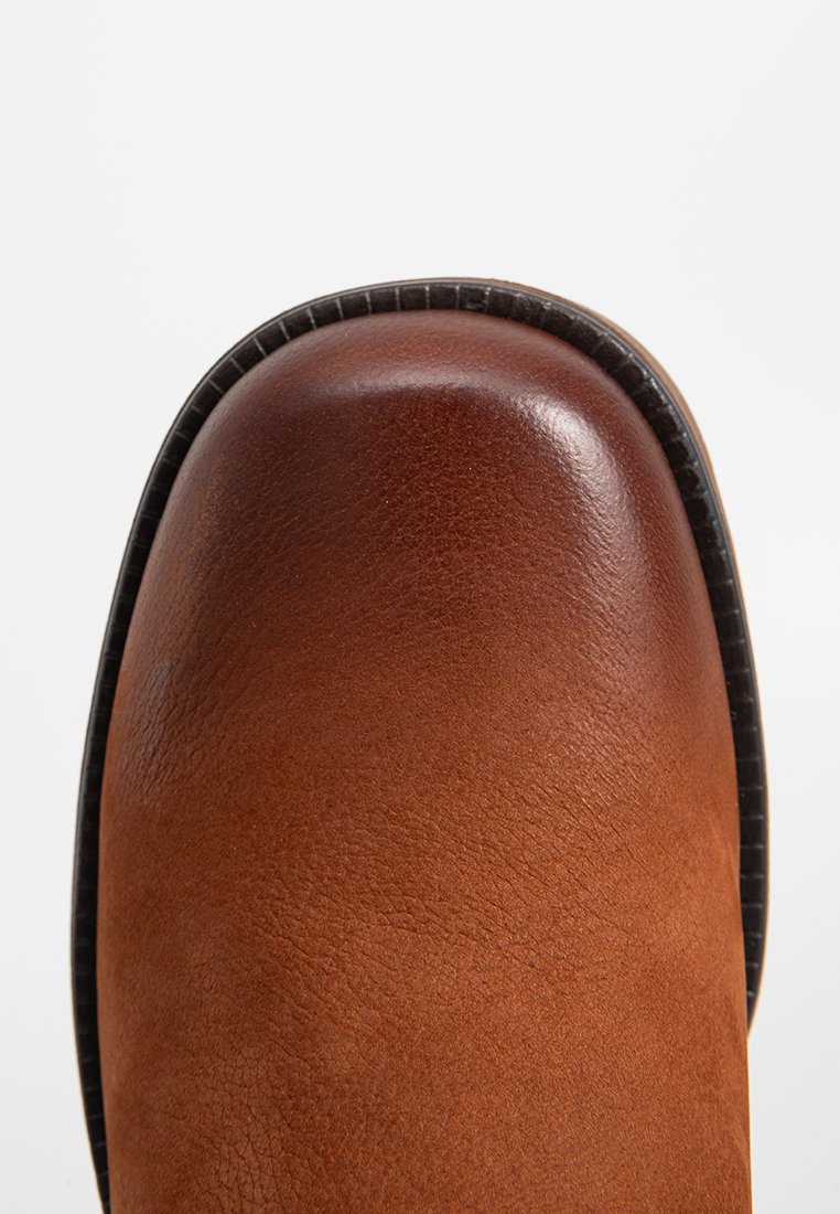 Bota de cuero marrón con un acabado suave, puntera redondeada y ribete de suela negra. La textura es ligeramente granulada, mostrando el grano natural del cuero.