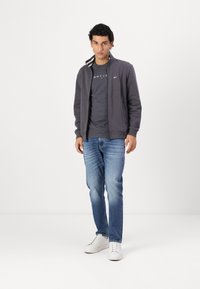 Giacca grigia con zip e colletto a righe, indossata sopra una maglia grigia scura. Abbinata a jeans azzurri chiari e scarpe bianche su uno sfondo neutro.