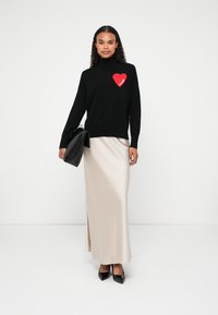Žena v čiernom svetri s červeným srdcom a textom "FUTURE", béžovej saténovej sukni, čiernych lodičkách a nesúca čiernu kabelku na bielom pozadí.