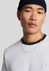 Witte T-shirt met een zwarte geribbelde kraag, met een klein lichtblauw geborduurd vogellogo op de borst, gedragen met een zilveren ketting en een marineblauwe beanie.