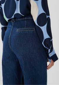 Jeans med hög midja i mörkblå denim, med en standard bakficka med diskret sömnad. Kombineras med en blå och marinblå blus med polka prickar.