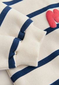 Sudadera a rayas en color crema con franjas azul marino y azul claro. Detalles: puños acanalados, letra "M" texturizada en rosa en la parte delantera.