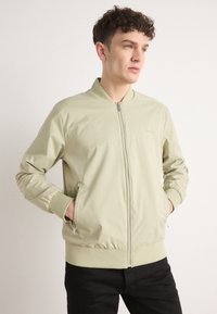 Hellgrüne Bomberjacke mit Reißverschluss, gerippten Bündchen und Saum, Seitentaschen und glatter, glänzender Textur. Minimale Markenkennzeichnung auf der Brust.