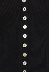 Černý žebrovaný cardigan s vertikálním pruhem kulatých, světlých knoflíků s malými otvory, který zdůrazňuje texturu a design.