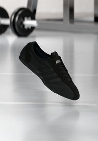 Μαύρο αθλητικό παπούτσι με λείο συνθετικό επάνω μέρος, κλείσιμο με κορδόνια, σχεδιασμό με τρεις ρίγες στο πλάι και στρογγυλεμένη μύτη.