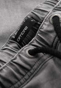 Pantalones cortos de tela gris con una cinturilla fruncida, que cuentan con un cordón negro y una etiqueta de tela que muestra el nombre de la marca "ombre."