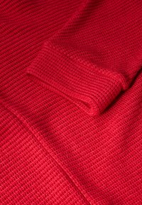 Maglione rosso lavorato a maglia con una superficie texturizzata. Ingrandimento del polsino della manica che mostra un design arrotolato e dettagli a costine.