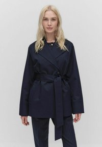 Granatowa kurtka zapinana na pasek o dopasowanym kroju, z szerokimi klapami i kieszeniami bocznymi; wykonana z gładkiego, lekkiego materiału.