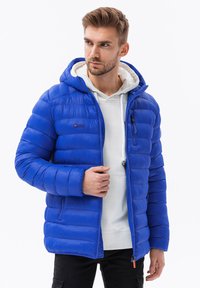 Giacca a vento blu con cappuccio, design trapuntato, chiusura con zipper e tasche laterali. Presenta un rivestimento interno bianco e dettagli arancioni sulla zip.