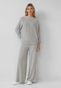 Frau, die gegen eine graue Wand steht und einen lockeren grauen Pullover, weit geschnittene graue Hosen und weiße Sneaker trägt.