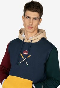 Felpa con cappuccio caratterizzata da un corpo blu navy, maniche bordeaux e verde, tasca gialla e motivo sportivo ricamato con mazze incrociate e cappellino.