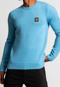 Man i en figursydd ljusblå stickad tröja med ribbade manschetter och nederkant, försedda med en liten svart och guld logotyp på bröstet.