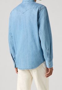 Jasnoniebieska koszula jeansowa z ostrym karczkiem, długimi rękawami i mankietami zapinanymi na guziki. Tkanina ma gładką fakturę i swobodny design.