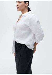 Chemise en lin blanc avec col et détails de boutons, associée à une jupe noire taille haute avec un lien latéral et des accents de franges.