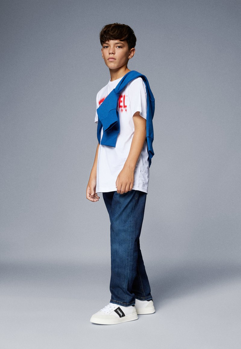 T-shirt bianca con testo rosso, maglione blu appoggiato sulle spalle, jeans blu scuro e sneakers bianche con accenti neri. Sfondo grigio.