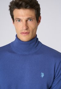 Maglione a collo alto blu realizzato in morbido materiale a maglia, con colletto a coste e un piccolo logo accentato sul lato sinistro del petto.