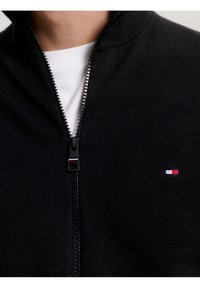 Svart zip-up tröja delvis åtdragen över en vit skjorta, med en liten röd, vit och blå rektangulär logotyp på bröstet.