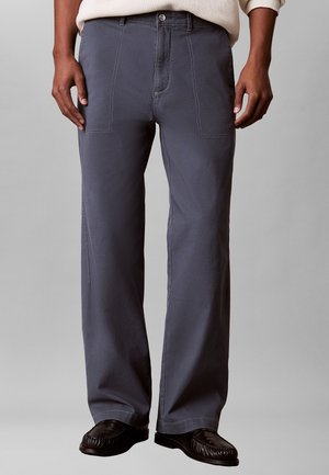 Man draagt grijze rechte broek met grote voorzakken, een lichte trui en zwarte leren schoenen tegen een effen achtergrond.