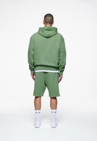 Sudadera verde con capucha y cordón, combinada con pantalones cortos a juego. Textura suave, corte relajado y detalles blancos en la cinturilla y los calcetines.