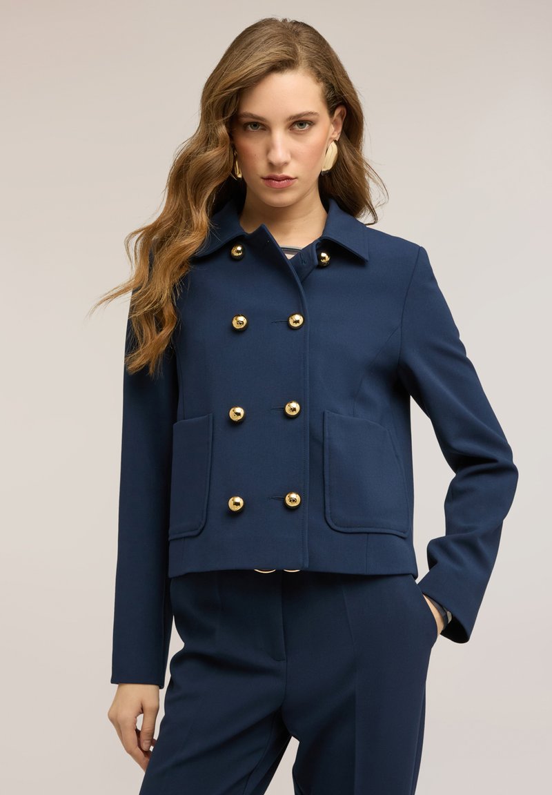 Mujer con cabello largo y ondulado, vistiendo una chaqueta azul marino de doble botonadura con botones dorados y pantalones a juego, de pie con una mano en el bolsillo.