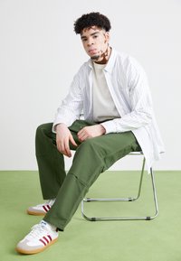 Gestreepte witte en blauwe shirt, beige sweatshirt, groene cargobroek en witte sneakers met rode accenten, zittend op een chroomstoel op groene vloer.