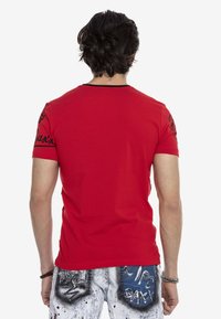 Rotes T-Shirt mit kurzen Ärmeln, schwarzem Ausschnitt und Logodetails an den Ärmeln, kombiniert mit grauen, zerrissenen Jeans mit künstlerischen Mustern und Akzenten.