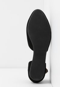 Semelle de chaussure plate noire tournée vers l'extérieur, chaussure positionnée verticalement contre un mur blanc avec un angle.