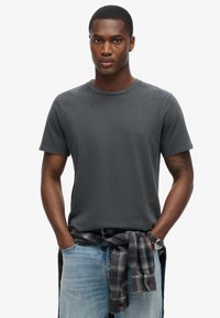 Mørkegrå kortærmet t-shirt med rund hals. Båret med lyseblå jeans og en ternet skjorte bundet om taljen. Har et ur på håndleddet.