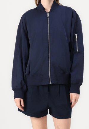 Person trägt eine marineblaue Bomberjacke mit Reißverschluss, gerippten Bündchen und Saum, kombiniert mit passenden marineblauen Shorts.