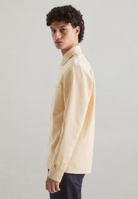 Light yellow button-up shirt with a textured fabric, featuring a chest pocket and long sleeves, viewed from the side against a neutral background.