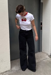 Bijela majica s crvenim ovratnikom i vezom trešnje, uparena s crnim trapericama širokih nogavica visokog struka. Pozicija stojeći s rukama na struku.