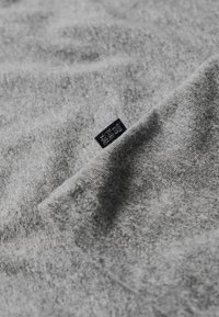 Grijze stoffen textuur met zachte plooien, met een klein donker label in de hoek, dat het merk of verzorgingsinstructies aangeeft.