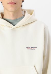 Sudadera con capucha en color crema, fabricada con tejido suave. Presenta un bolsillo frontal y un logo bordado en azul y rojo. Superpuesta sobre una camiseta verde.