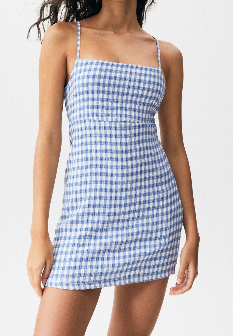 Femme portant une mini-robe à carreaux bleus et blancs, sans manches, avec de fines bretelles, sur un fond clair uni.