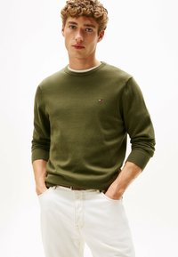 Maglione lavorato a maglia verde oliva con scollatura a girocollo, polsini a coste e piccolo logo in evidenza. Abbinato a pantaloni chiari per creare contrasto.