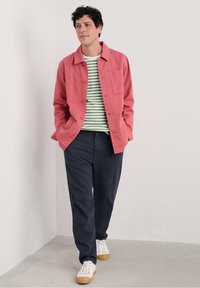 Veste en coton corail avec fermeture à boutons, associée à une chemise rayée verte et blanche, un pantalon bleu marine et des baskets blanches avec des semelles en gomme.