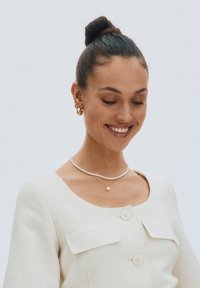 Weiß strukturierte Jacke mit zwei vorderen Taschen, getragen über einer Perlenkette und einer zarten Kette mit einem kleinen Perlenanhänger.