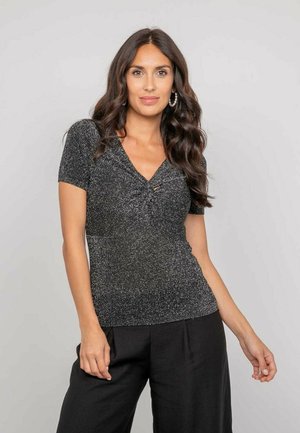 Femme aux longs cheveux ondulés portant un haut noir scintillant à manches courtes et un pantalon noir, posant devant un fond gris clair uni.