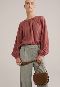 Rūsgana blūze ar savākta kakla izgriezumu un teksturētu rakstu, apvienota ar augsto jostasvietu pelēkiem haļšiem ar šaha rakstu un brūnu zamša rokassomu.