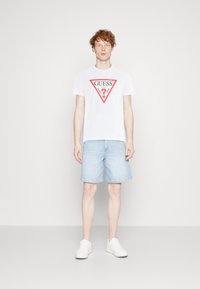 White short-sleeve t-shirt featuring a red triangular logo with "GUESS USA" text. Paired with light blue denim shorts and white sneakers.