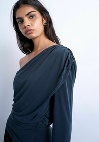 Femme aux longs cheveux noirs portant une robe asymétrique bleu foncé et des boucles d'oreilles géométriques dorées sur un fond clair uni.