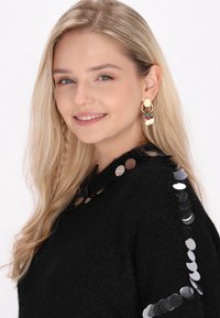 Svart stickad tröja med paljetter på axlarna. Modellen bär färgglada, flerlagrade örhängen med runda, metalliska inslag.