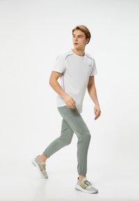 Witte t-shirt met korte mouwen en grijze accentstiksels, gecombineerd met lichtgroene joggingbroeken en grijze sneakers met gele en zilveren accenten.