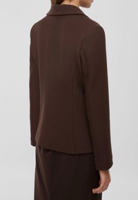Blazer marrone a texture con colletto, vestibilità sartoriale, silhouette dritta e maniche lunghe. La vista posteriore mostra i dettagli delle cuciture e linee pulite.