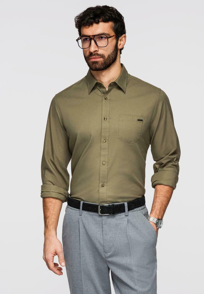 Homme aux cheveux bouclés foncés et à la barbe portant des lunettes à monture écaille de tortue, une chemise vert olive boutonnée avec les manches roulées, un pantalon gris et une montre.