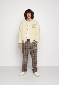 Cream shearling bomber jacket with black and white striped cuffs, brown plaid pants with embroidered detail, and chunky white sneakers.