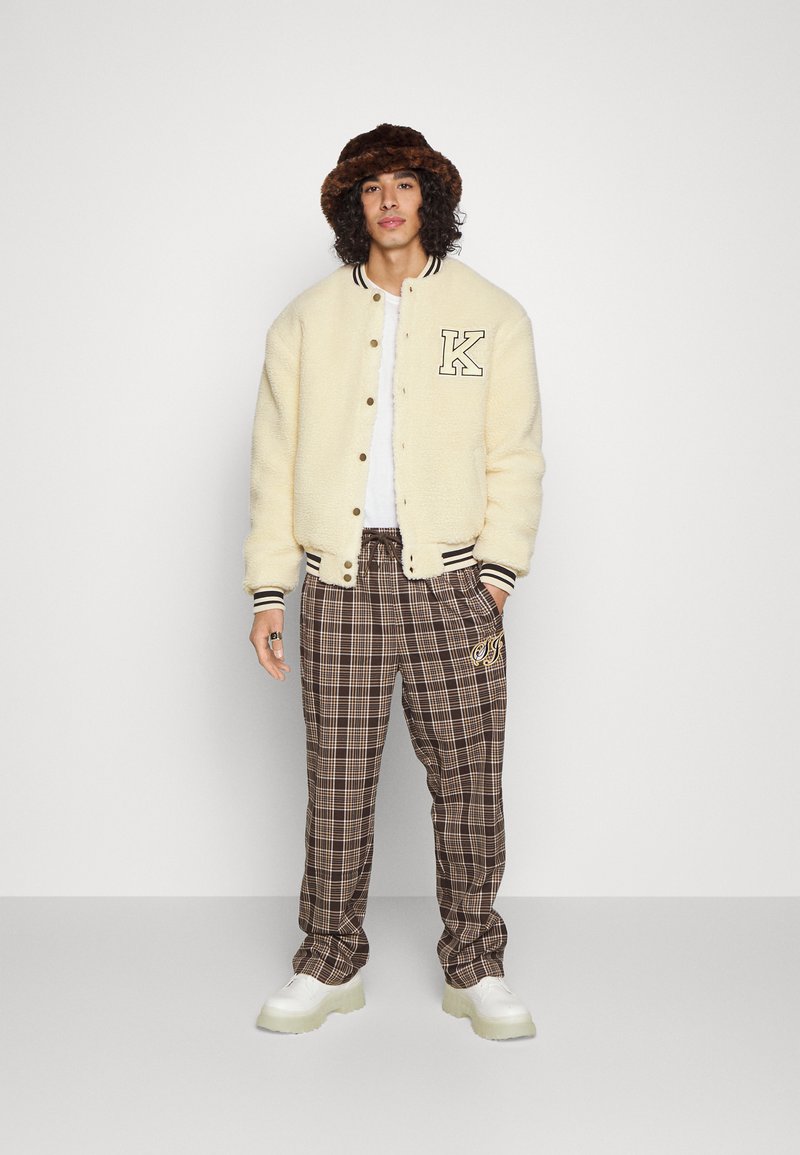 Chaqueta bomber de borreguillo crema con puños a rayas en blanco y negro, pantalones de cuadros marrones con detalles bordados y zapatillas chunky blancas.